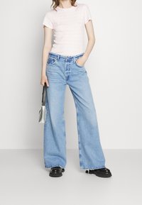 Rosa randig t-shirt med kort ärm, matchad med vida ljusblå jeans. Modellen håller en liten metallicklindad handväska och bär svarta kraftiga skor.