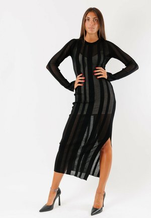 Donna che indossa un lungo vestito nero trasparente a righe con maniche lunghe e tacchi alti neri, in piedi con le mani sui fianchi su uno sfondo bianco.
