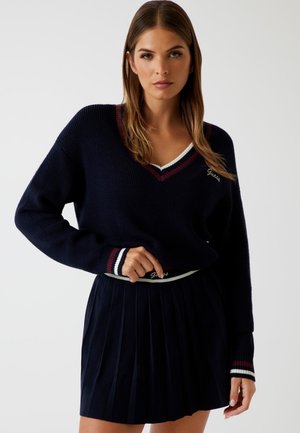 Marineblauer Strickpullover mit V-Ausschnitt und kontrastierendem burgunderrot-weißen Besatz; kombiniert mit einem plissierten marineblauen Rock mit einem markierten Bund.