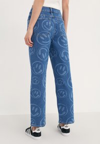 Vaqueros de mezclilla en azul con un patrón de caras sonrientes repetido, diseño de pierna recta, cintura media y bolsillos traseros. Combinados con zapatillas negras y blancas.