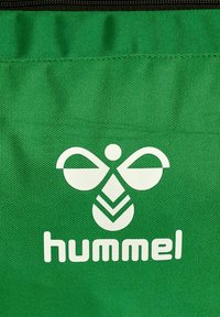 Bolsa de tela verde con un logo blanco que presenta formas abstractas y la palabra "hummel" en la parte inferior. Superficie texturizada con cierre de cremallera.