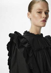 Schwarze Bluse mit Rüschen und gewellten Ärmeln sowie einem plissierten Design. Der Stoff wirkt leicht und hat eine glatte Textur.
