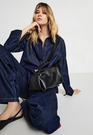 Borsa nera in pelle con forma strutturata, accenti dorati e una tracolla removibile, appoggiata su un modello in abbigliamento in denim scuro.
