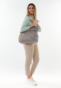 Donna con maglione verde menta e pantaloni beige che porta una grande borsa a tracolla grigia con cerniere gialle, in piedi su uno sfondo bianco.