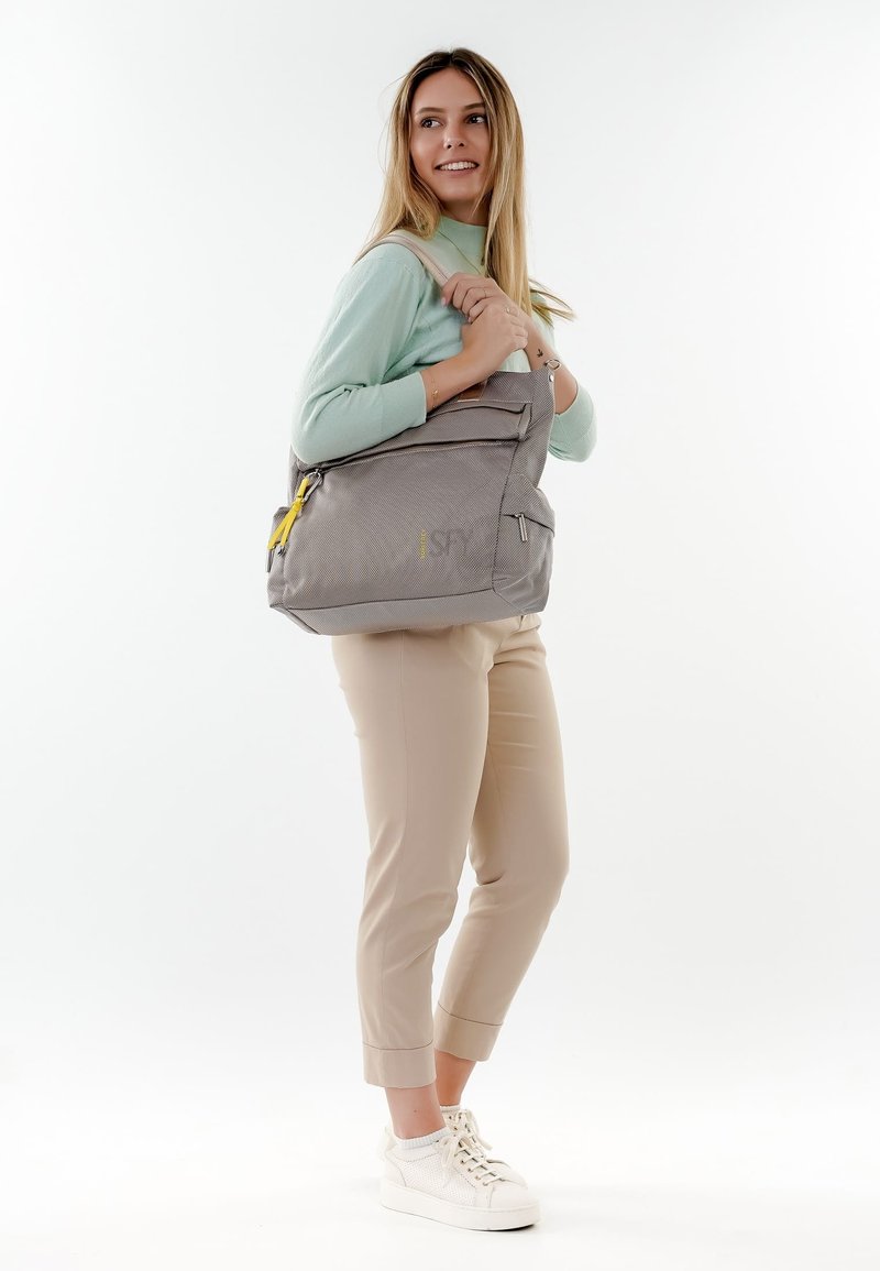 Donna con maglione verde menta e pantaloni beige che porta una grande borsa a tracolla grigia con cerniere gialle, in piedi su uno sfondo bianco.