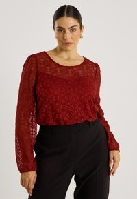 Blouse rouge en dentelle avec manches longues et motif ondulé texturé, dotée d'un col rond. Associée à un pantalon noir taille haute.