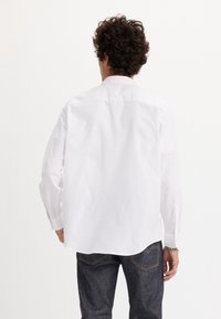 Muž s kudrnatými vlasy oblečený v bílém tričku s dlouhým rukávem a tmavě modrých džínách, stojící zády k fotoaparátu.