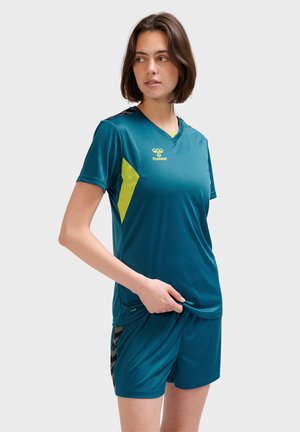 Młoda kobieta ubrana w turkusowy strój sportowy z żółtymi detalami stoi i patrzy na bok na jednolitym tle.