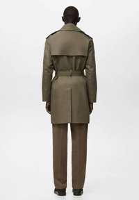 Gabardina caqui con cintura abrochada y hombreras, con abertura en la parte trasera. Combinada con pantalones marrones y zapatos negros.