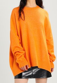 Un gros pull orange vif en matériau doux. Il a un col rond, des épaules tombantes et un ourlet côtelé. Associé à une jupe noire brillante.