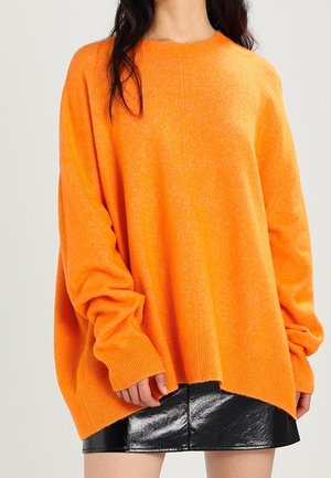 Maglione oversize arancione brillante realizzato in materiale morbido. Presenta un collo rotondo, spalle scese e orlo a costine. Abbinato a una gonna nera lucida.
