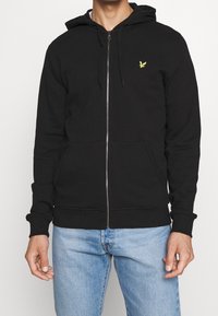Sudadera con cremallera negra en mezcla de algodón, con un bolsillo tipo canguro, puños de canalé y un pequeño logo amarillo en el pecho. Textura suave.
