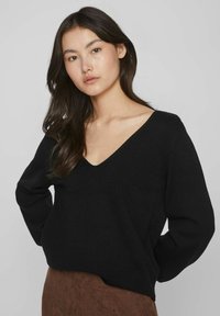 Suéter negro de cuello en V en un suave tejido de punto con mangas largas, que presenta un corte relajado y dobladillo acanalado, combinado con una falda marrón texturizada.