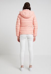 Różowa kurtka puchowa z kapturem, z poziomym pikowaniem i elastycznymi mankietami, w zestawieniu z białymi obcisłymi jeansami i białymi trampkami.