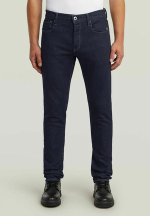 Slim fit donkerblauwe spijkerbroek gedragen met zwarte veterlaarzen en een wit overhemd, voorkant zichtbaar van taille tot voeten.