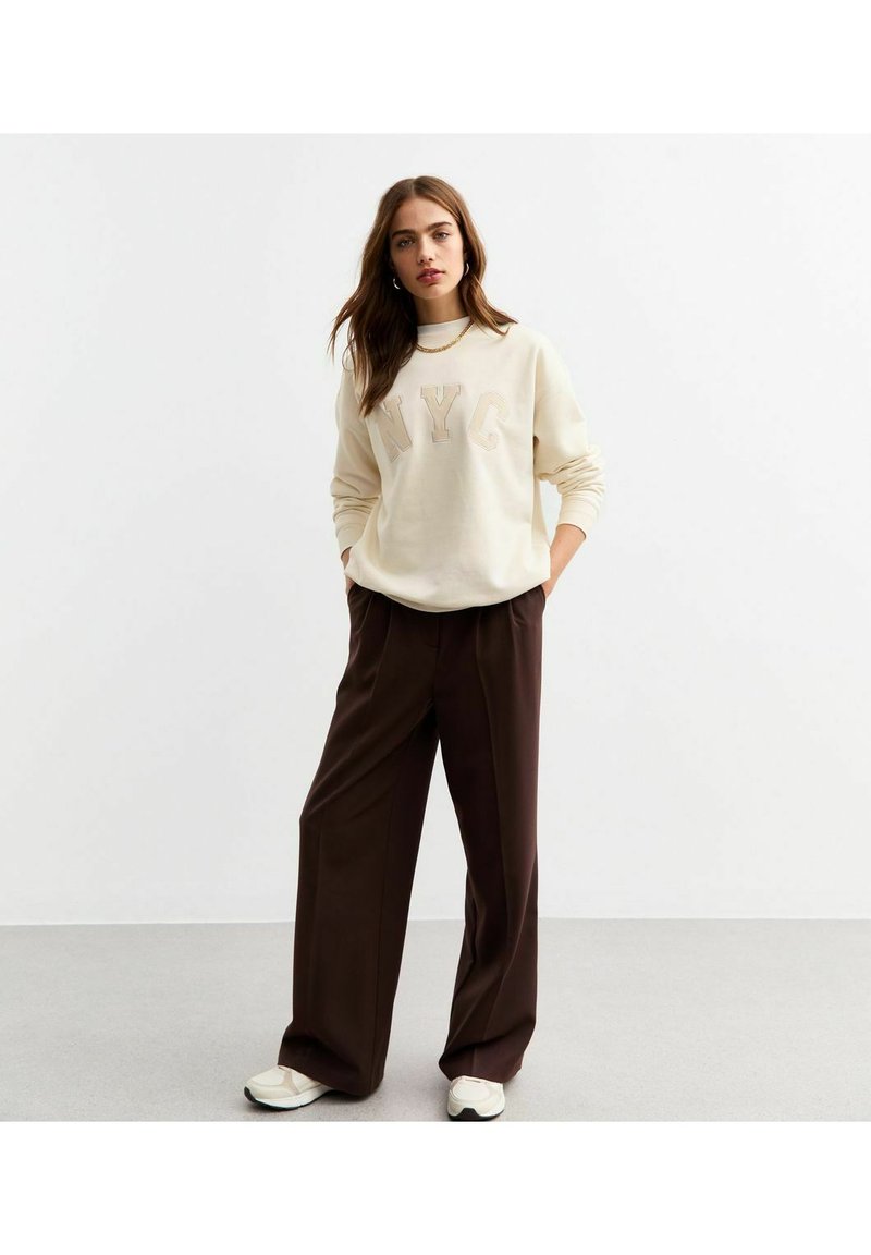 Cremefärgad sweatshirt med "NYC" i fet stil, kombinerad med lösa, högmidjade bruna byxor och vita sneakers. Släta tyger, avslappnad passform.