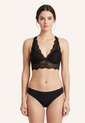 Žena v čiernom čipkovanom bralete s vlnitými okrajmi a zodpovedajúcim čiernym bezšvovým spodným prádlom, stojaca pred jednoduchým svetlým pozadím.