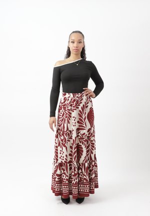 Mujer con una blusa negra de un solo hombro y manga larga y una falda blanca hasta el tobillo con estampado floral rojo, de pie con una mano en la cadera.