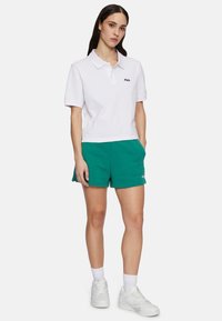 Vit kortärmad polo med krage och svart logotyp, kombinerad med gröna bomullsshorts. Vita sneakers och vita strumpor fulländar outfiten.