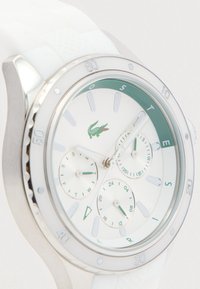 Montre Lacoste blanche avec trois sous-cadrans, lunette argentée marquée par des incréments et un logo crocodile vert sur le cadran.