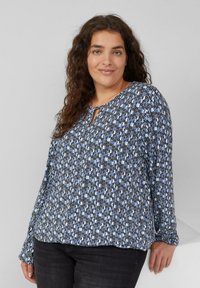 Schwarz-blau gemusterter Langarmoberteil mit einem Schlitz-Ausschnitt. Stoff aus weichem Material, der locker fällt. Enthält geometrische Formen in verschiedenen Größen.