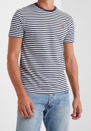 Homme portant un T-shirt à manches courtes à rayures horizontales bleu marine et blanc, ainsi qu'un jean bleu clair, se tenant devant un fond uni.