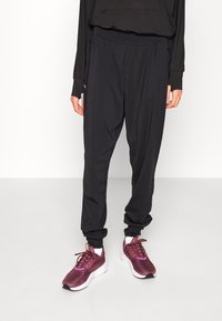 Schwarze Sporthose mit elastischem Bund, taillierter Passform und seitlichen Taschen, kombiniert mit burgunderfarbenen Sneakers mit weißen Sohlen.