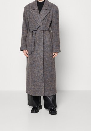 Długi szary wełniany płaszcz z paskiem związanym w talii, noszony na czarnych spodniach o szerokich nogawkach i czarnych butach, stojący na jednolitym białym tle.