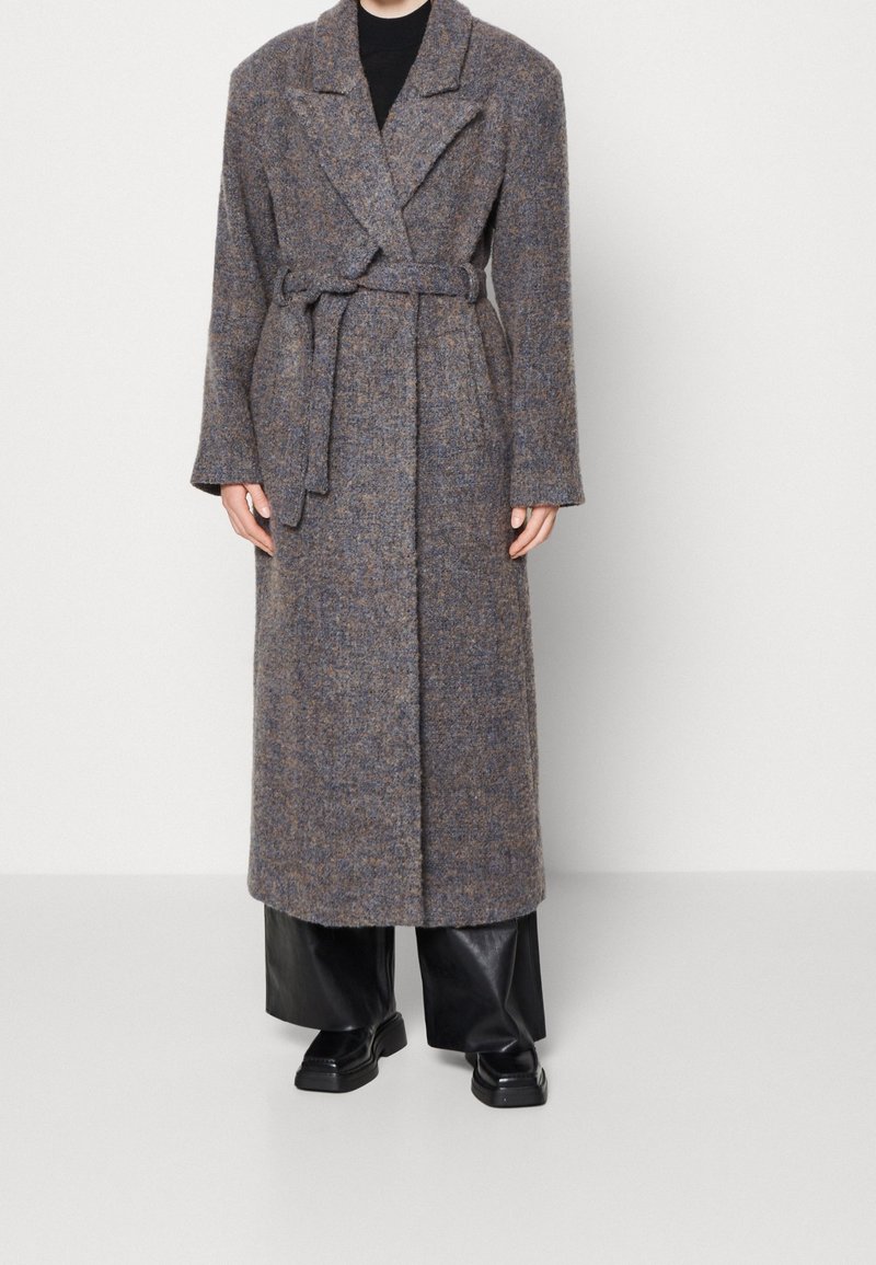 Long manteau en laine gris avec ceinture nouée à la taille, porté sur un pantalon large noir et des chaussures noires, debout devant un fond blanc uni.