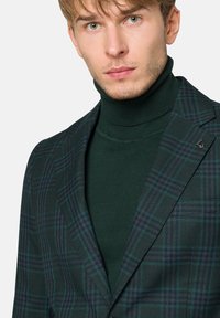 Zielona marynarka w kratę o wyraźnej linii, z ciemnoniebieskimi akcentami i z klapą, noszona na zielonym swetrze z golfem.