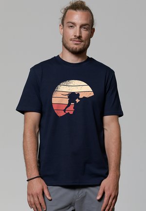 Mann trägt ein marineblaues Hemd mit einem kreisförmigen Sonnenuntergangs-Motiv, das die Silhouette eines Kletterers zeigt, der gegen orange und beige Streifen an einer Felswand klettert.