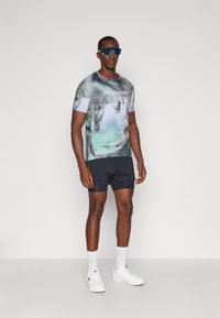 Buntes grafisches T-Shirt mit einem pixelierten Effekt, kombiniert mit dunklen Shorts. Die Accessoires bestehen aus reflexionsfähigen Sonnenbrillen und weißen Sneakers.