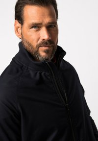 Marinblå zip-up jacka med ribbad krage och manschetter, som har en slät yta och strukturerad design. Dragkedjesmyckning är synlig.