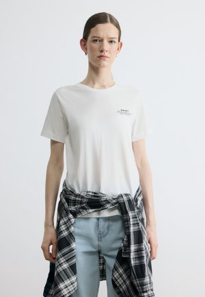 Giovane donna con indosso una t-shirt bianca GANT, jeans azzurri, con una camicia a quadri bianca e nera legata intorno alla vita, in piedi davanti a uno sfondo bianco.