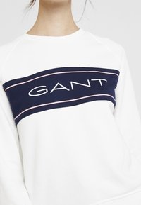 Witte sweatshirt met een marineblauwe horizontale band met het "GANT"-logo in het wit, geaccentueerd met een dunne roze lijn. Zacht materiaal, ronde hals.