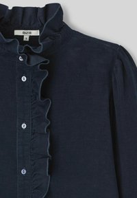 Chemise en velours côtelé bleu foncé avec un col froncé haut, une fermeture à boutons à l'avant et des manches légèrement bouffantes. Présente de petits boutons blancs.