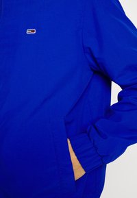 Blue windbreaker jacket with elastic cuffs and side pockets. Features a small logo patch on the left chest. Made from lightweight fabric.