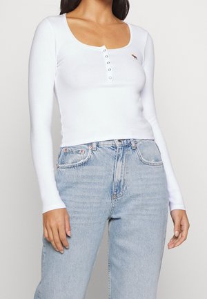 Weißes Langarmshirt mit Rundhals-Ausschnitt, versehen mit einer Knopfleiste und einem gestickten Detail auf der Brust, kombiniert mit hellblauen Jeans.