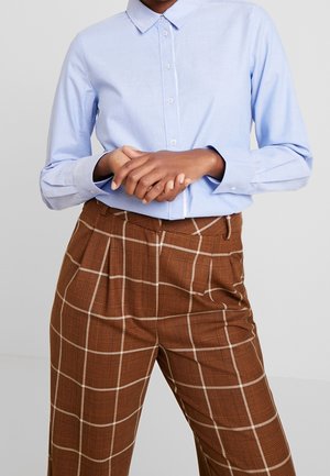 Persoon die een lichtblauw overhemd met knopen en een bruine geruite broek met hoge taille draagt, met de handen gevouwen voor zich, staand tegen een effen achtergrond.