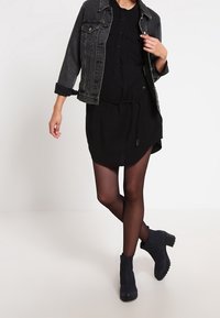 Vestido negro de camisa con cierre frontal de botones, ajustado en la cintura. Combinado con una chaqueta de mezclilla corta y botas oscuras hasta el tobillo.