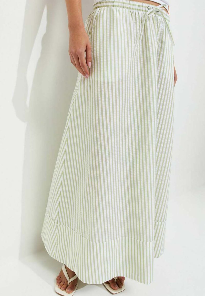 Woman wearing a long white and light green vertical striped skirt with elastic waistband and white flip-flops, standing against a white wall.