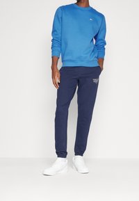 Sudadera azul con cuello redondo y puños acanalados, combinada con pantalones deportivos azul marino con texto de logo en blanco, complementada con zapatillas blancas.