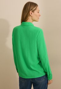 Blusa lunga a maniche lunghe di colore verde con texture, colletto, polsini con bottoni e vestibilità comoda, mostrata da dietro su uno sfondo neutro.