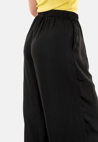 Pantalones negros fluidos con cintura elástica y bolsillos laterales. Textura suave y diseño de pierna ancha visible desde la vista lateral.