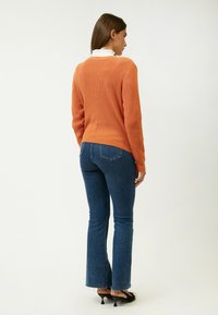 Orange ribbet sweater med en ensfarvet ryg, parret med blå folderede jeans. Sættet inkluderer sorte højhælede sko med en struktureret detalje.