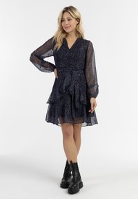 Tumma kukkamekko, jossa on teksturoitu, läpinäkyvä pitkähiha, kietaisumalli, kerrokselliset röyhelöt ja istuva vyötärö. Yhdistetty mustiin paksupohjaisiin saappaisiin.