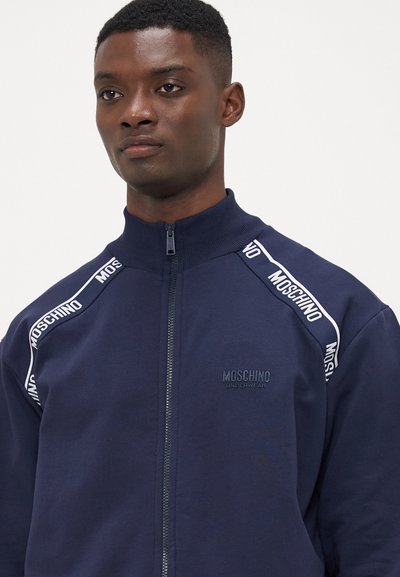 Mornarska zip-up jakna z visokim ovratnikom, ki ima belo logotipno trak ob ramenih in subtilen embosiran logotip blagovne znamke na prsih.