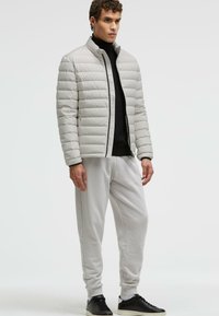 Hellgraue gepolsterte Jacke mit horizontalen Linien, schwarzem Reißverschluss, getragen über einem schwarzen Rollkragenpullover; kombiniert mit hellgrauen Jogginghosen und schwarzen Sneakers.