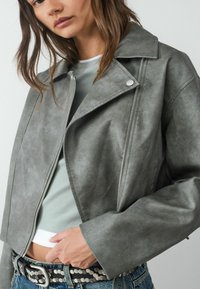 Grijze faux leren cropped jas met een ingezette kraag, zilveren hardware en een gladde textuur. Gewaard over een lichtgroene top.