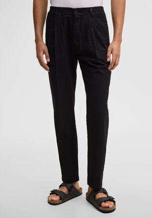 Mann trägt schwarze, plissierte Hosen und schwarze Sandalen mit Schnalle, steht vor einem schlichten hellen Hintergrund.